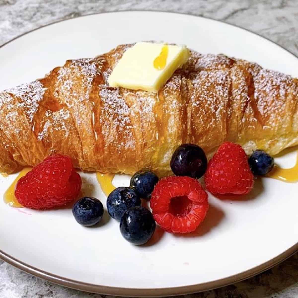 Croissant on plate topped with butter powdered sugar and honey sitting beside fruit