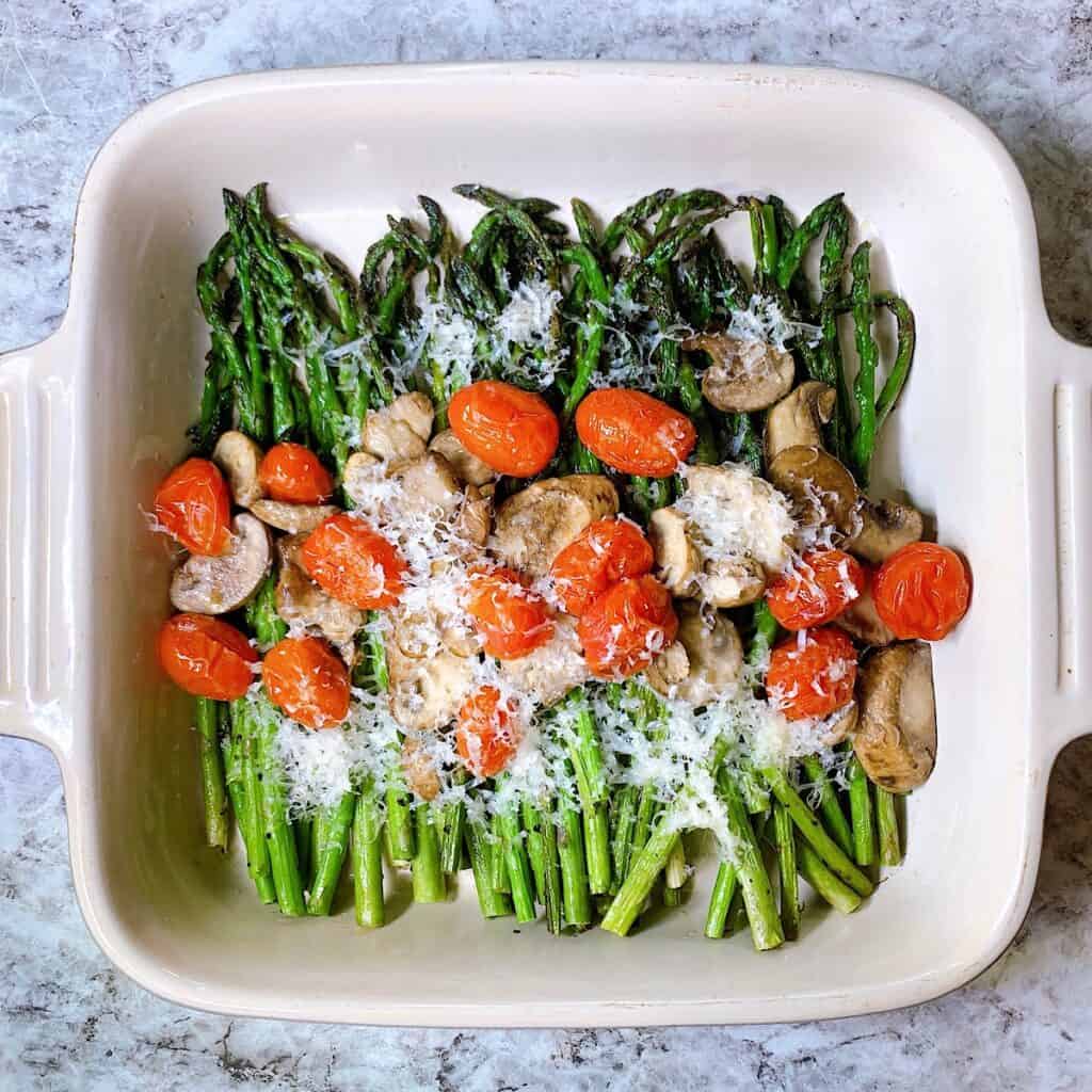 A casserole dish filled with roasted asparagus and topped with tomatoes and mushrooms