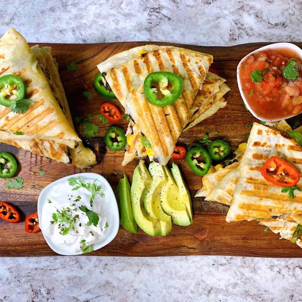 Sliced Chicken Quesadillas on a cutting board with bowls of sour cream salsa and garnished with cilantro avocado and jalapeno