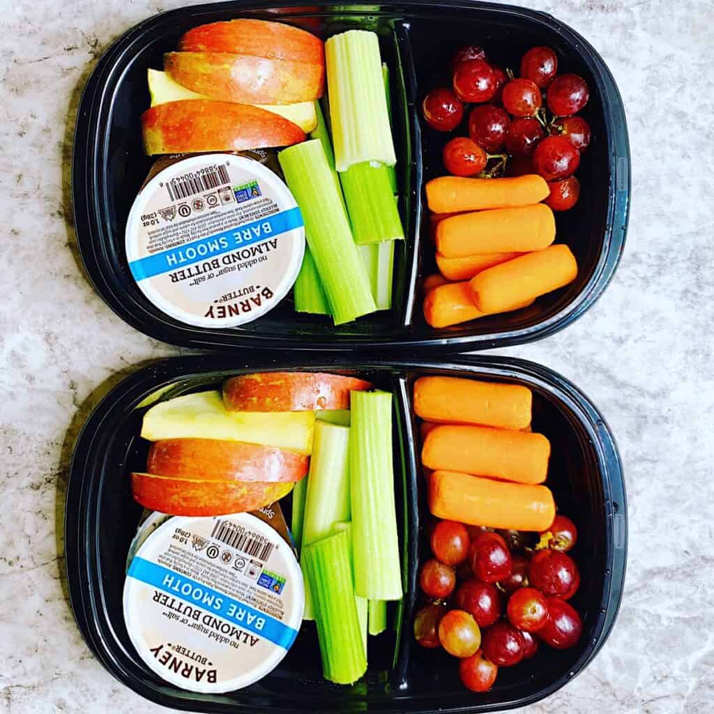 Meal prep containers with carrots, red grapes, sliced apple, celery, and barney butter almond butter cups.