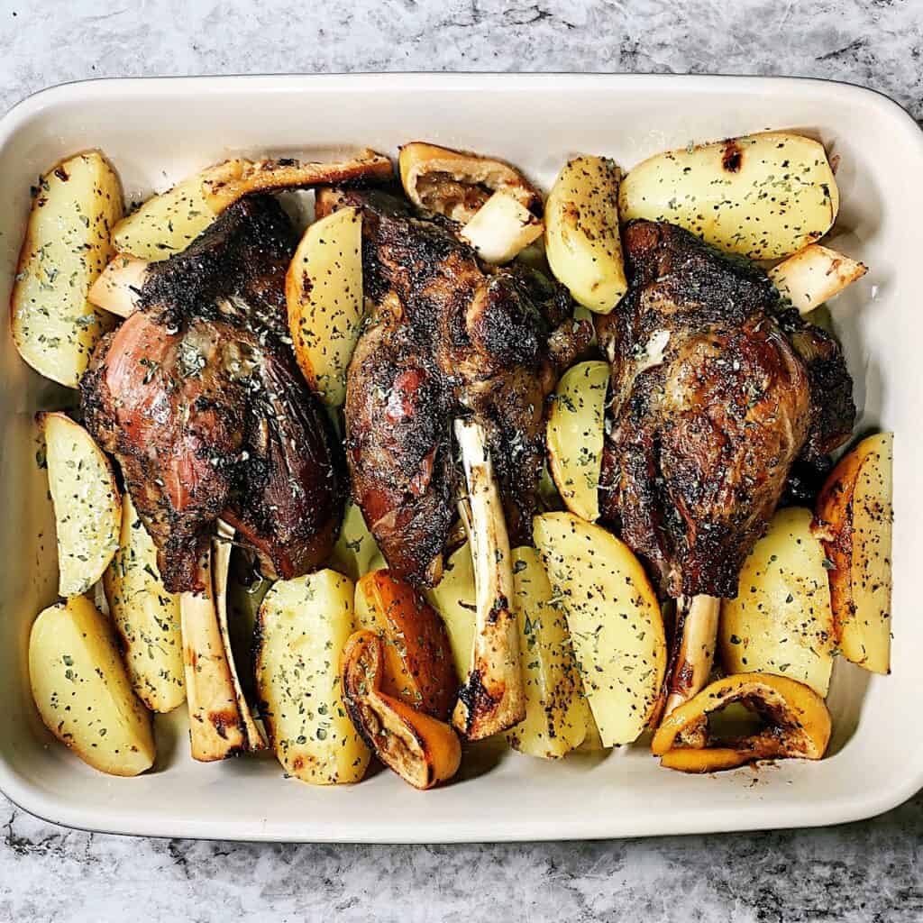 Roasted lamb shanks nestled between Greek potatoes in casserole dish