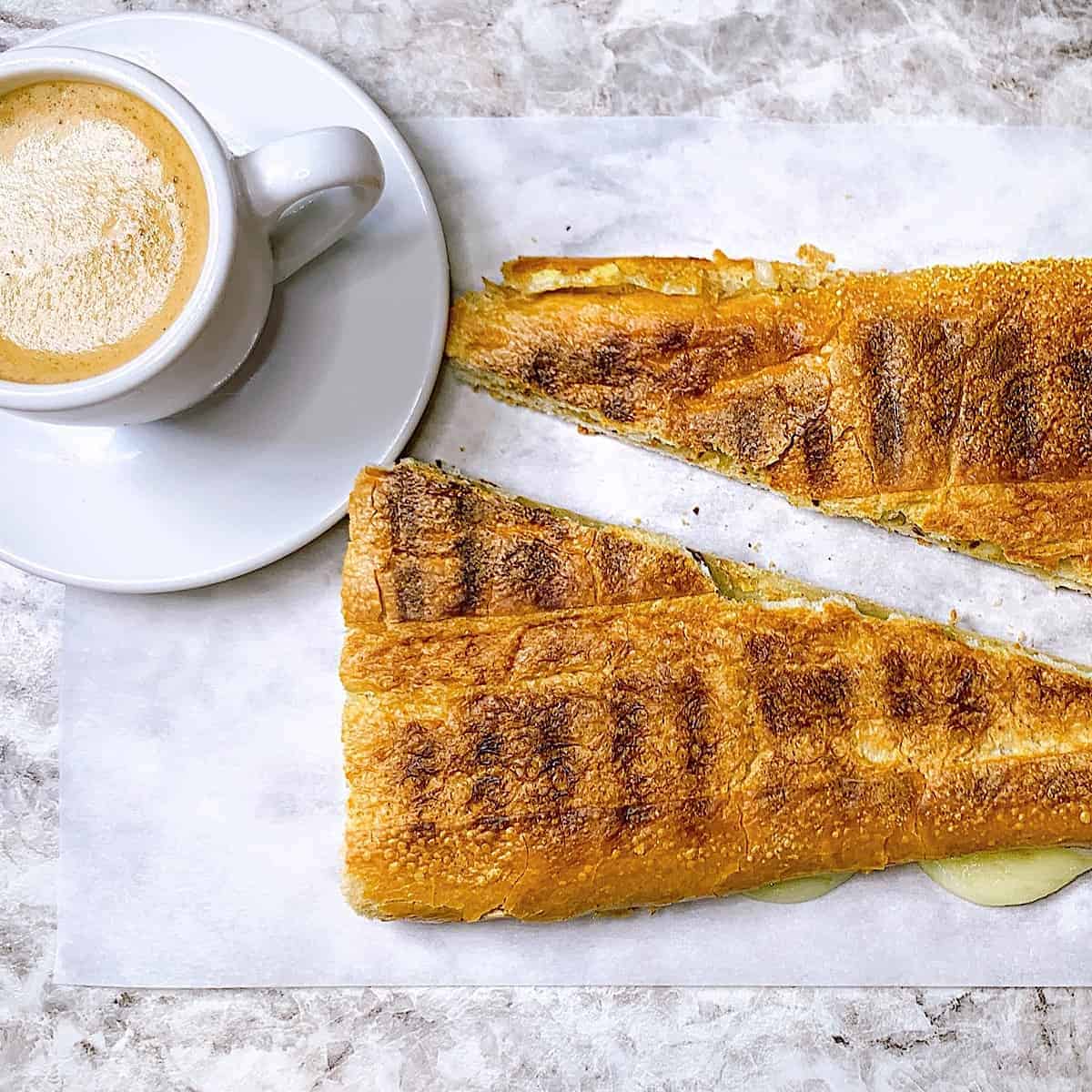 A small white cup of espresso and cream on saucer placed next to a large sliced cheesy pressed cuban bread