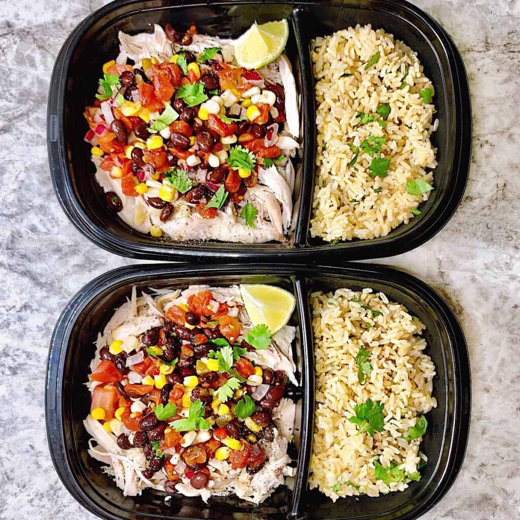 Two meal prep containers with rotisserie chicken, cilantro brown rice and topped with pico de gallo, black beans, corn, cilantro, and lime.