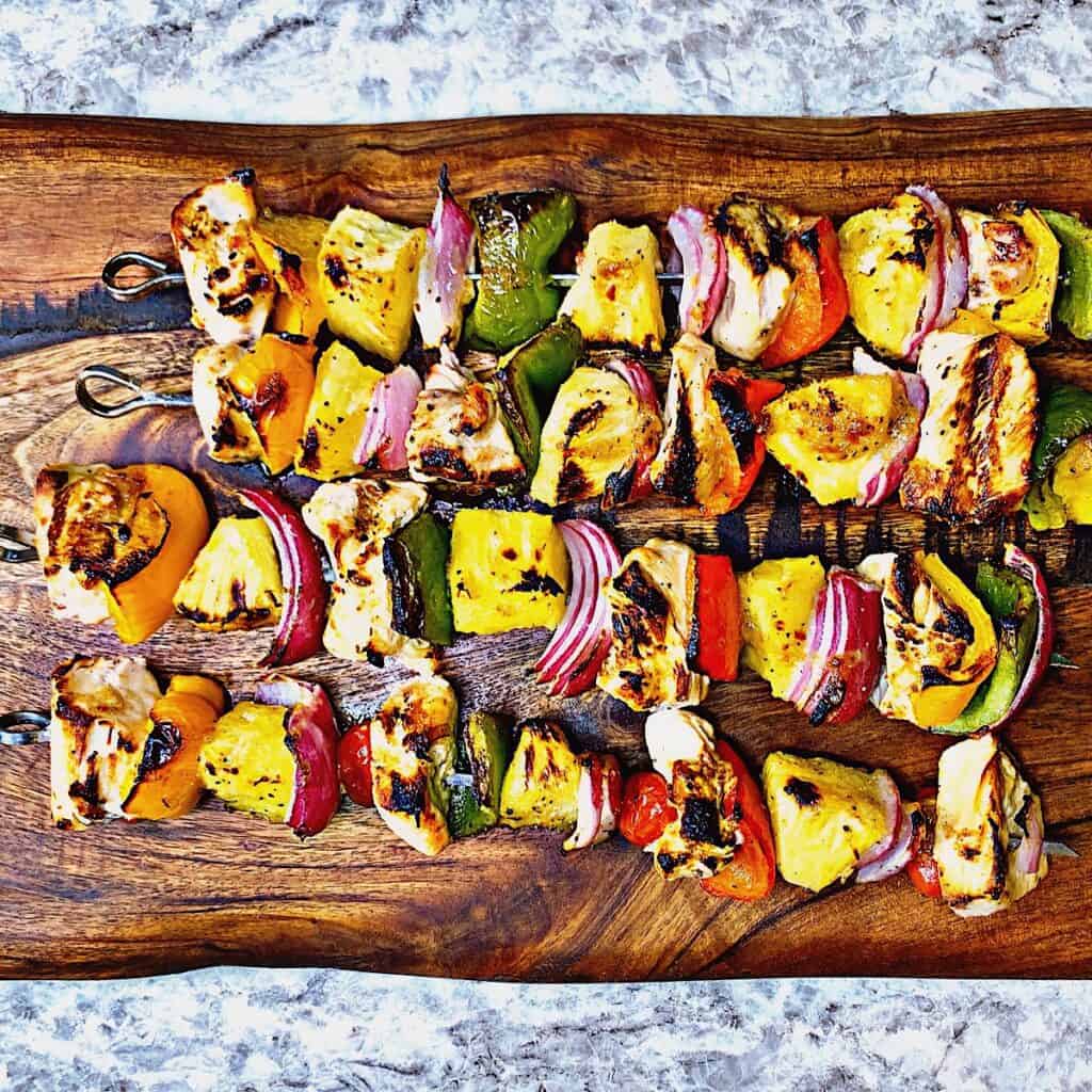 Charred grilled chicken kabobs with peppers, onion, and pineapple on metal skewers displayed on wooden cutting board