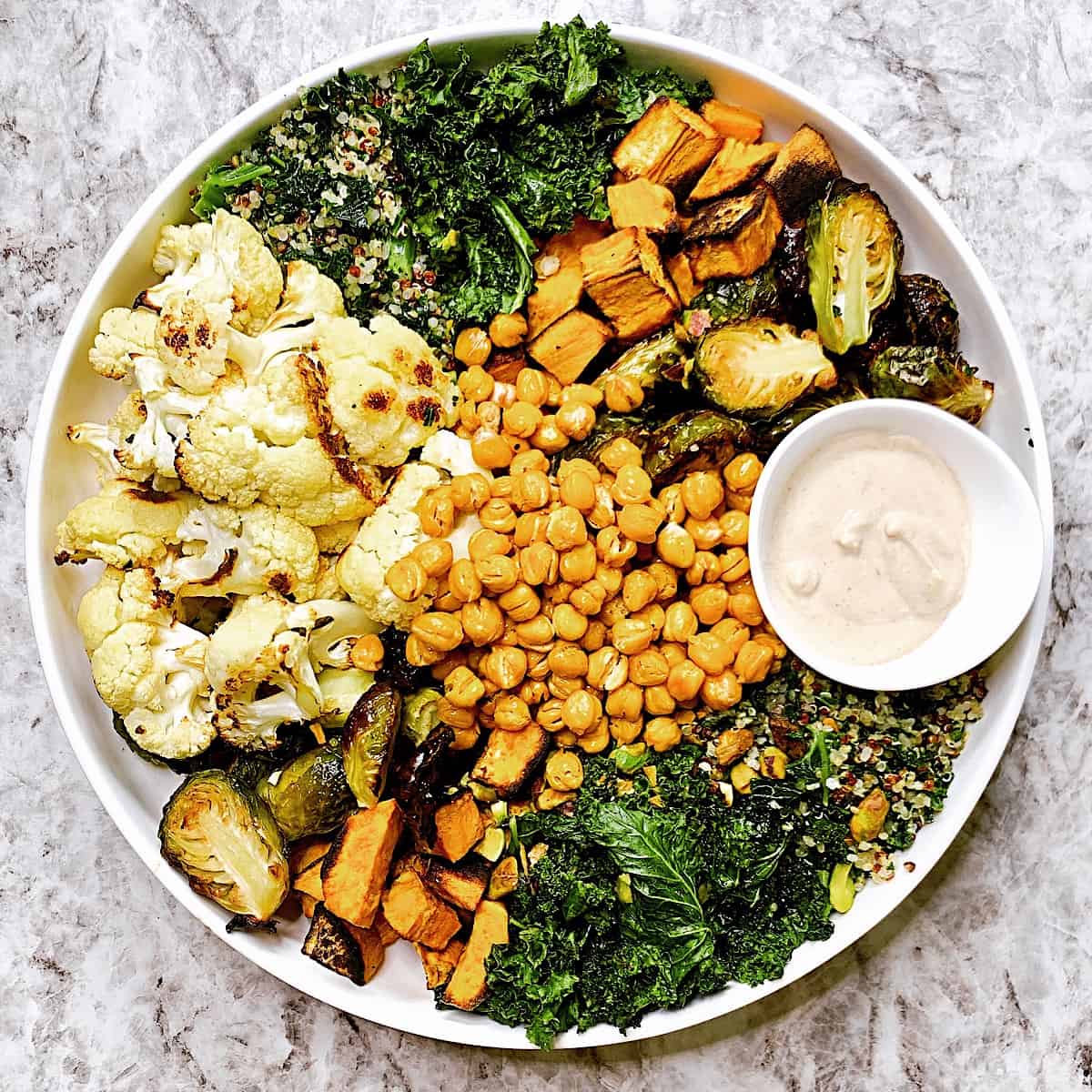 Large bowl filled with roasted kale, cauliflower, sweet potatoes, brusselsprouts, chickpeas, and quinoa with tahini sauce