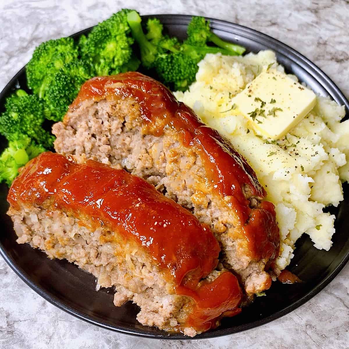 Sliced meatloaf on a black plate with mashed potatoes and broccoli