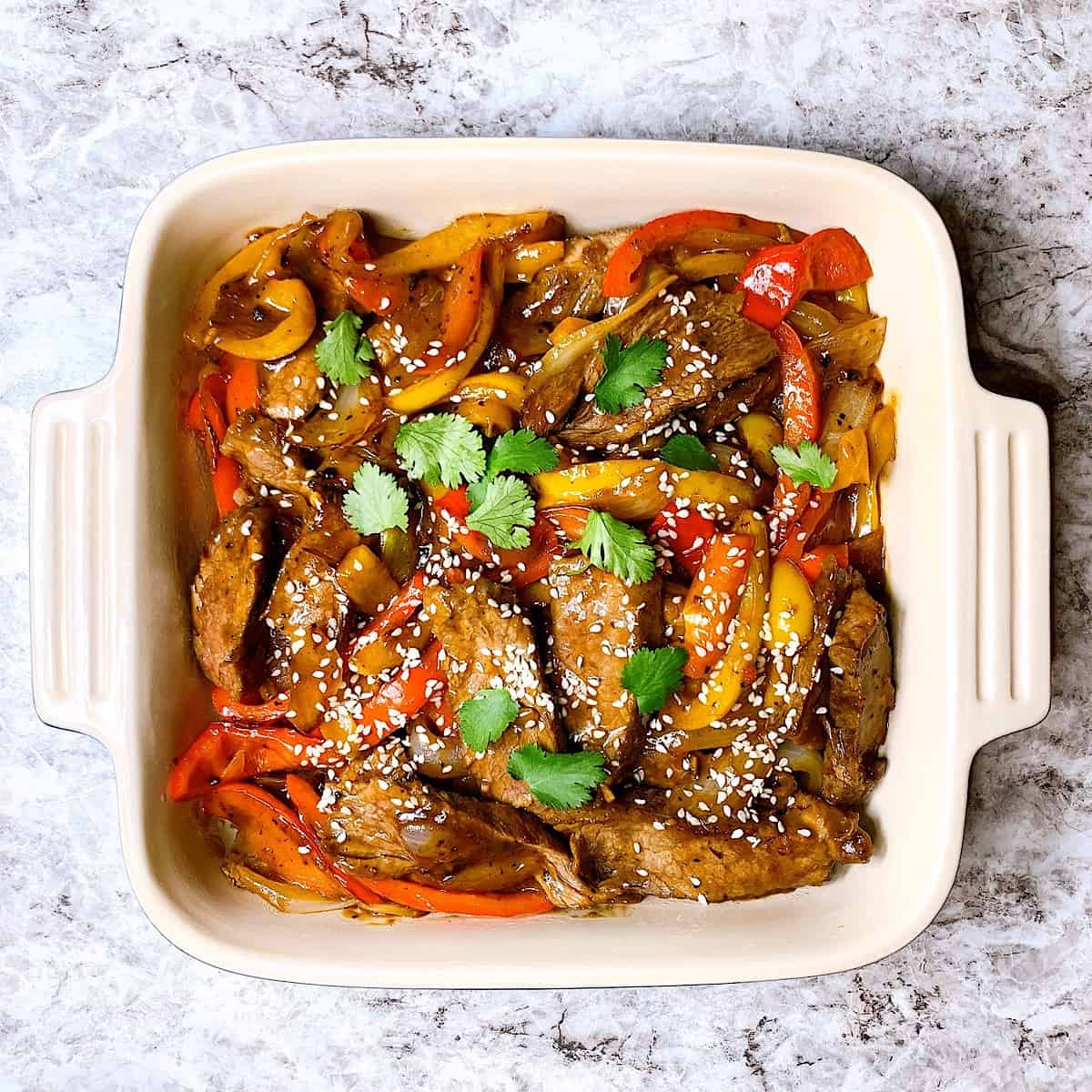 Pepper steak topped with yellow and red peppers cilantro and sesame seeds in casserole dish