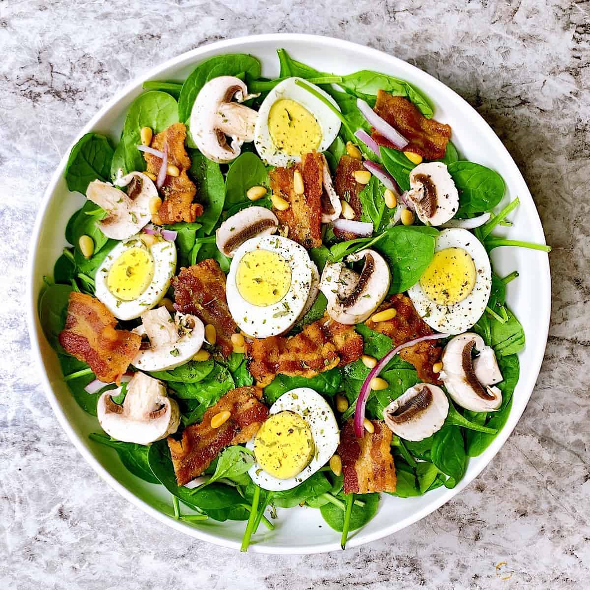 Spinach salad in large white bowl topped with bacon mushrooms and eggs