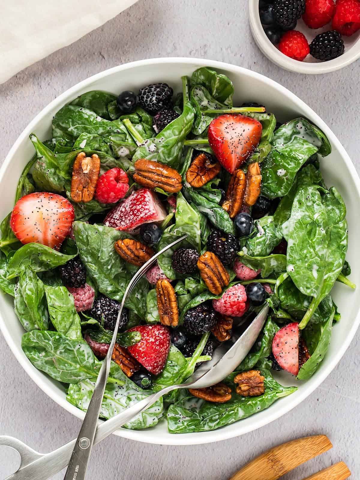 A berry spinach salad in a white serving bowl with linen and a small bowl of berries nearby.