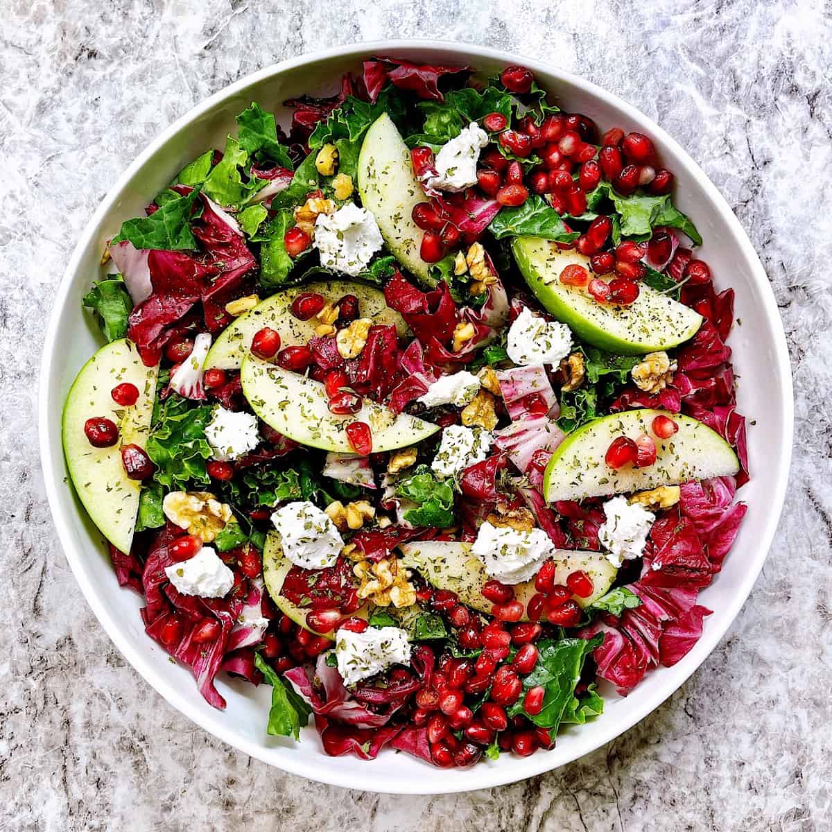 Apple and Pomegranate Salad in white bowl topped with walnuts and goat cheese