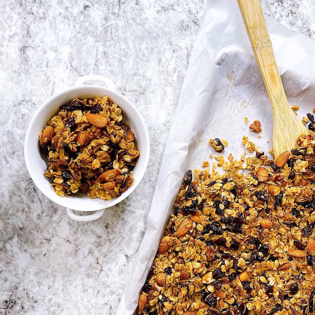 Cranberry Orange Granola on sheet pan with wooden spoon next to a bowl full of granola