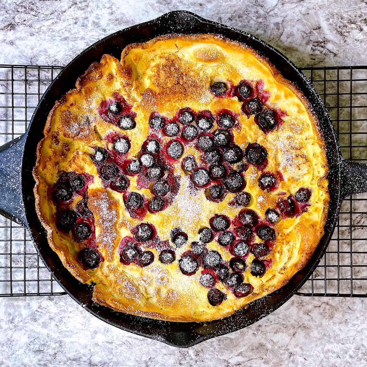 Dutch Baby Pancakes in cast iron skillet on baker’s rack with a sprinkle of powdered sugar and blueberries