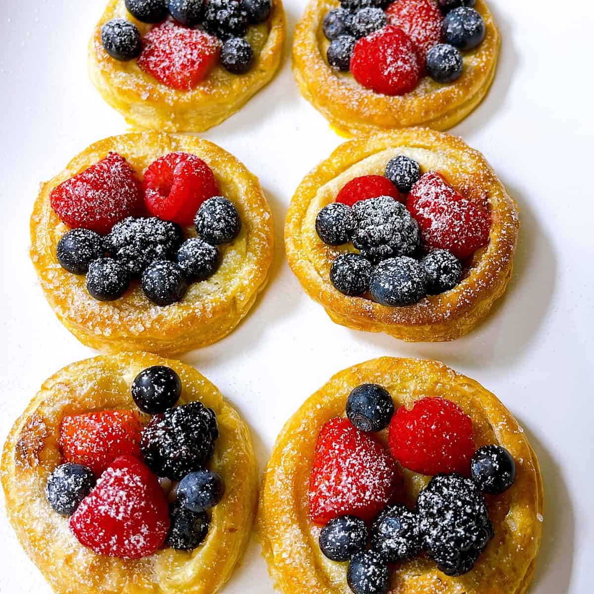 Mini Cheese Puff Danishes​ with powdered sugar on a white platter and topped with fruit