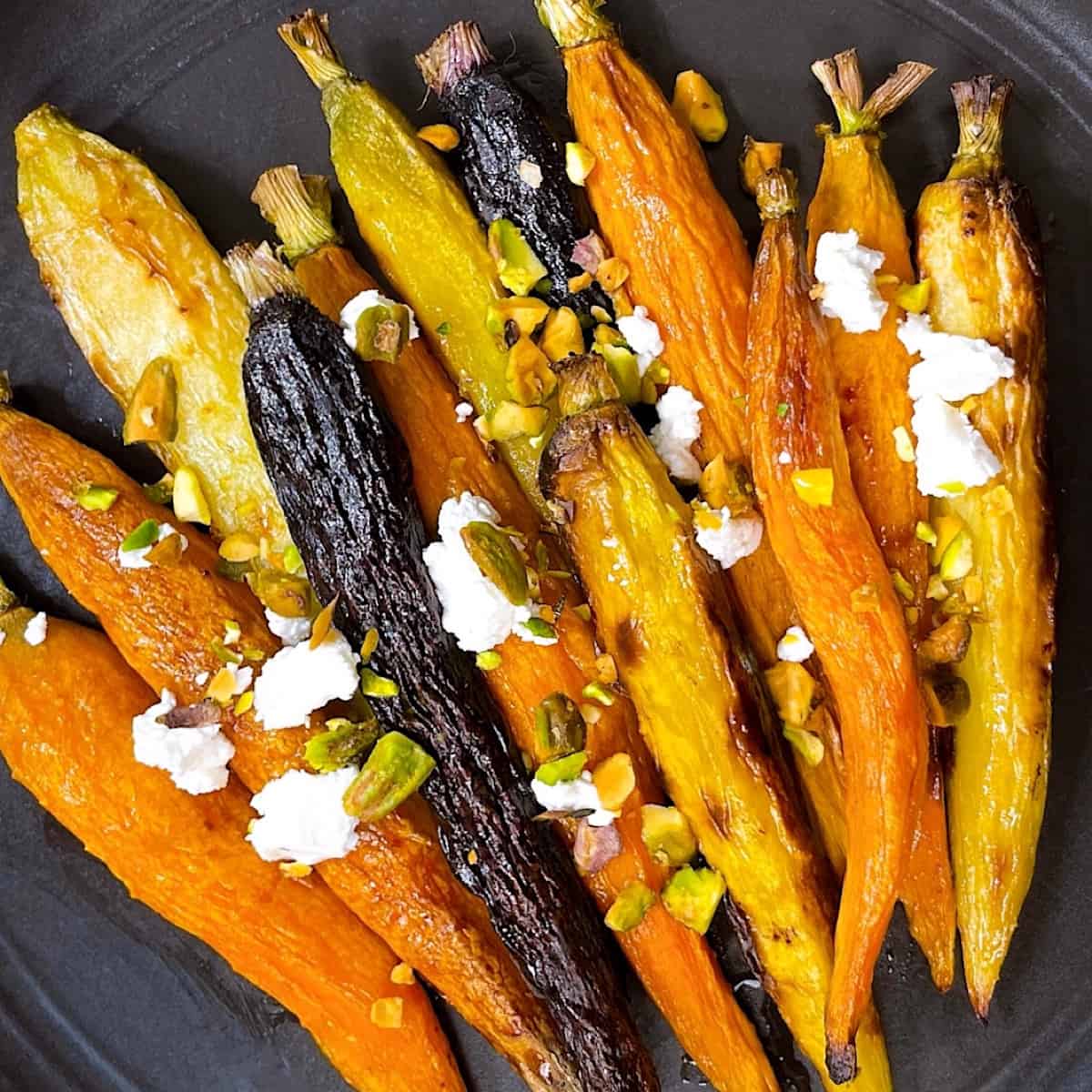 Roasted Rainbow Carrots on black plate topped with goat cheese, crushed pistachio, and drizzled with honey