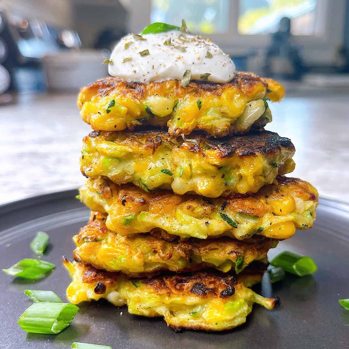 Zucchini and Corn Fritters on black plate with sliced green onions and a dollop of sour cream