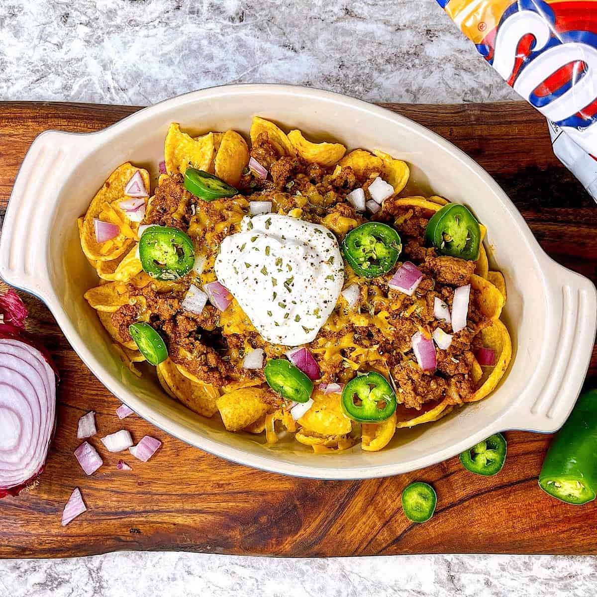 Frito Pie in casserole dish topped with onion jalapeño and sour cream sitting next to Frito bag
