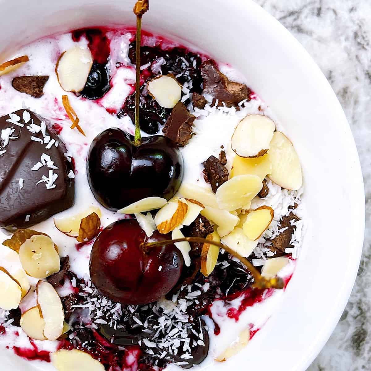 Warm Cherry Compote over Vanilla Ice Cream in white bowl topped with almonds chocolate and coconut