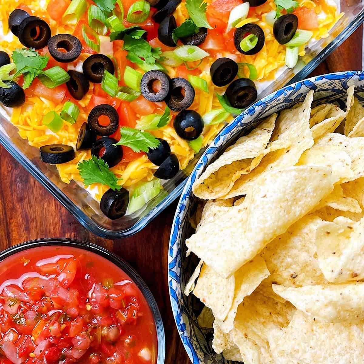 Seven Layer Dip next to a bowl of chips and salsa on wooden cutting board