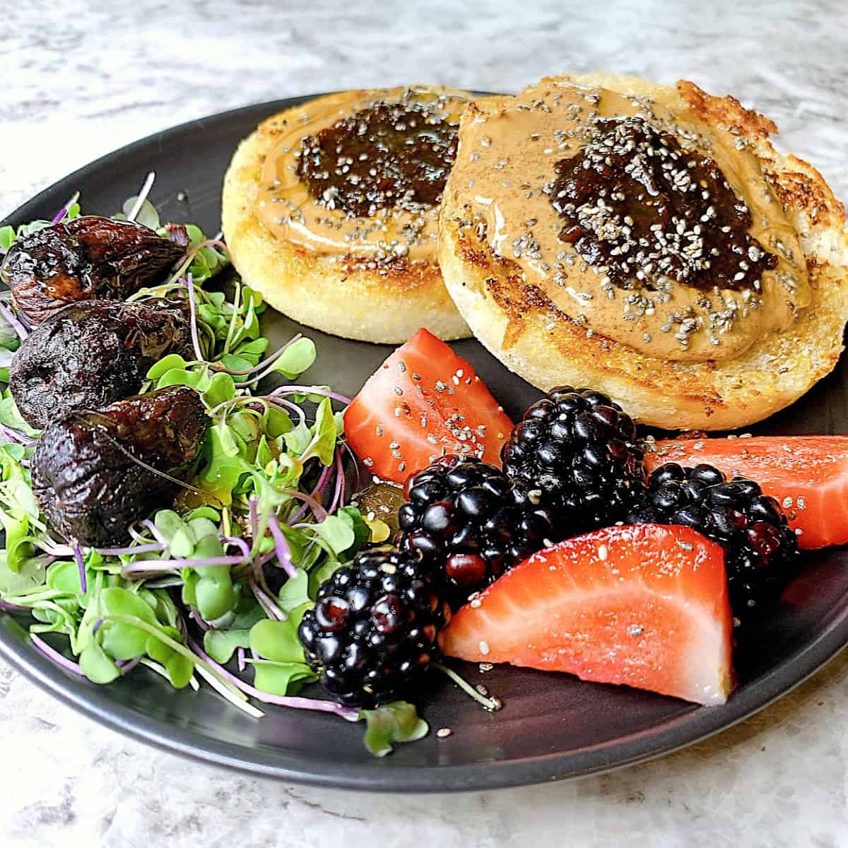 Toasted english muffin topped with almond butter and fig jelly plated with microgreens and berries on a black plate