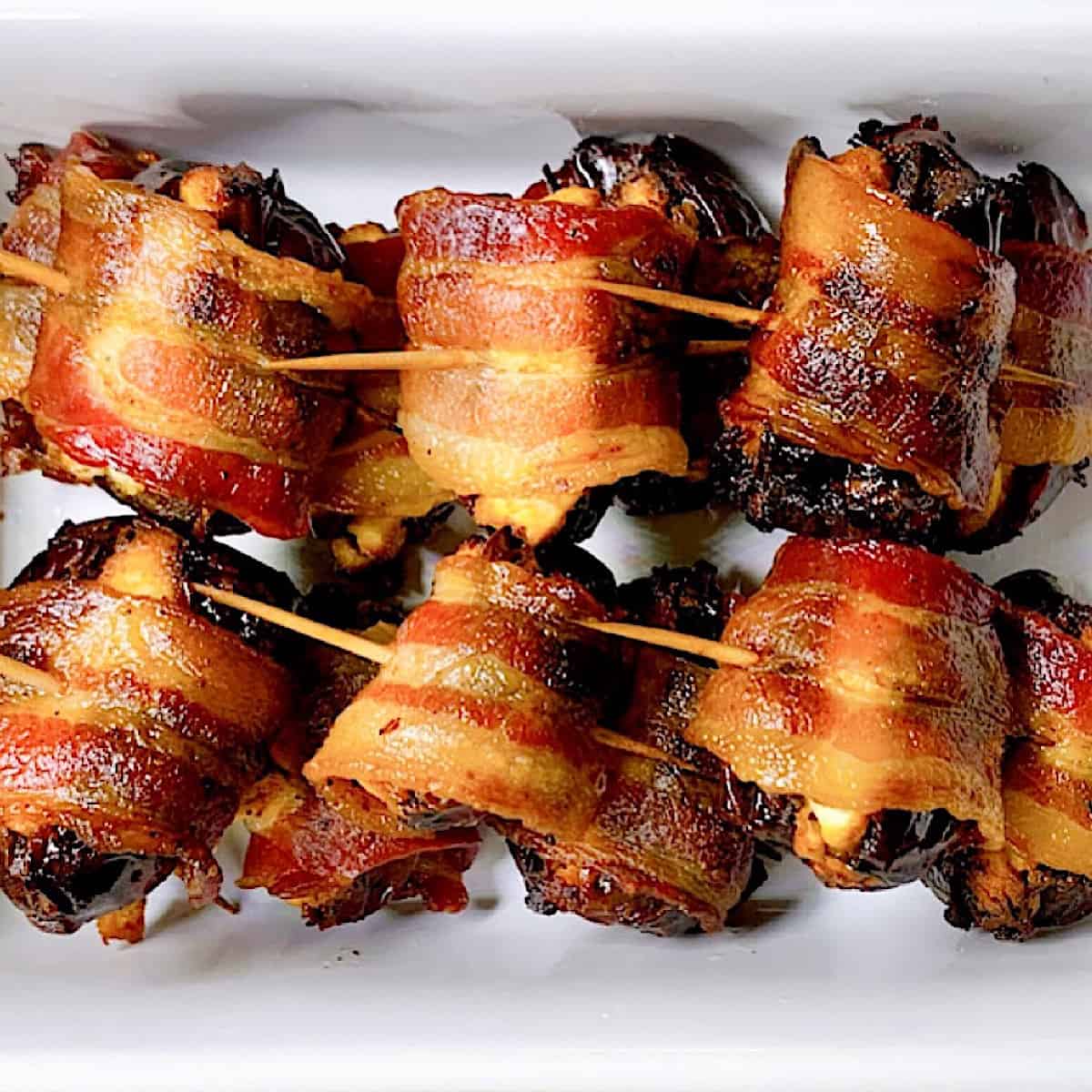 Six Bacon wrapped dates with toothpicks in small white ceramic crock close up
