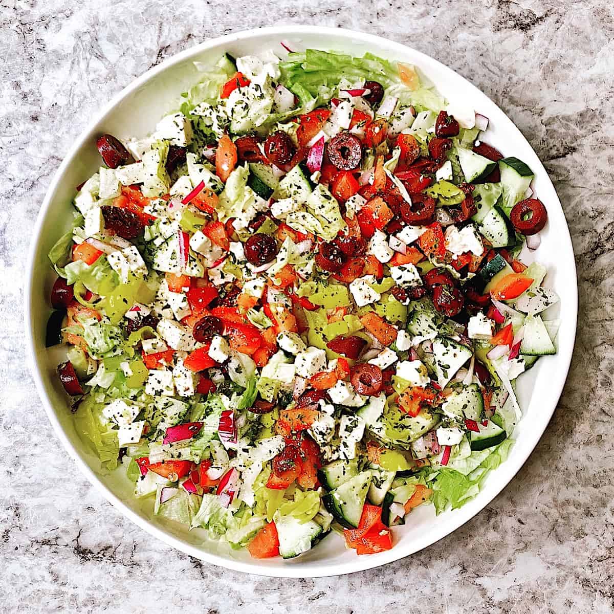 A large white bowl filled with chopped salad that includes feta, tomatoes, greek olives, greek peppers, cucumber, and oregano sprinkled on top.