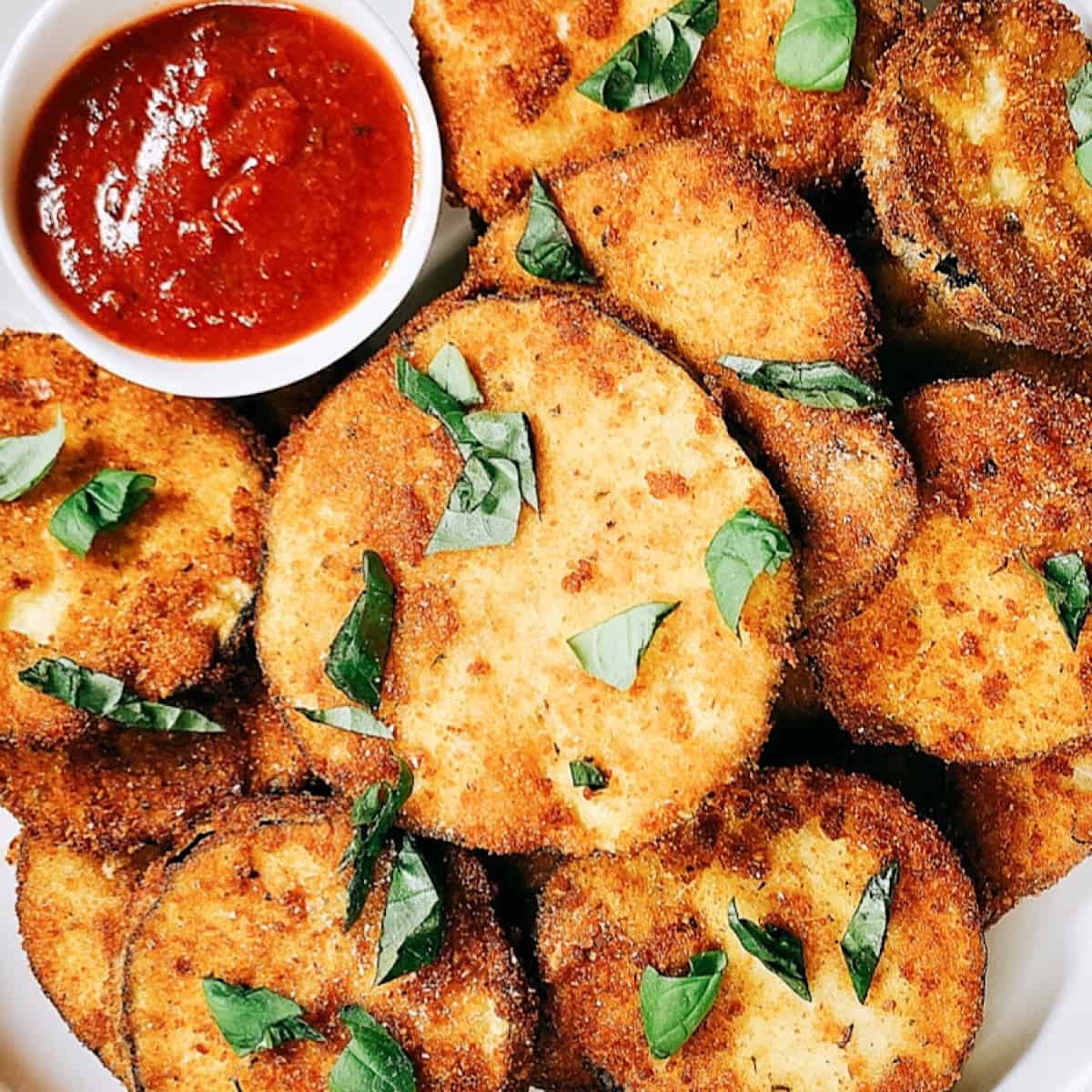 Fried eggplant in large white bowl with a side of marinara for dipped and topped with basil