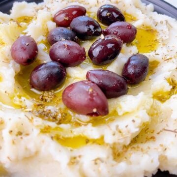 Skordalia Greek Potato dip with garlic in a black bowl topped with kalamata olives and extra virgin olive oil.