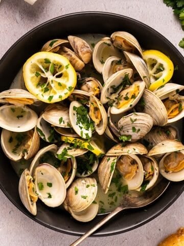 Easy steamed littleneck clams in a black bowl plated next to linen, parsley, and bread.