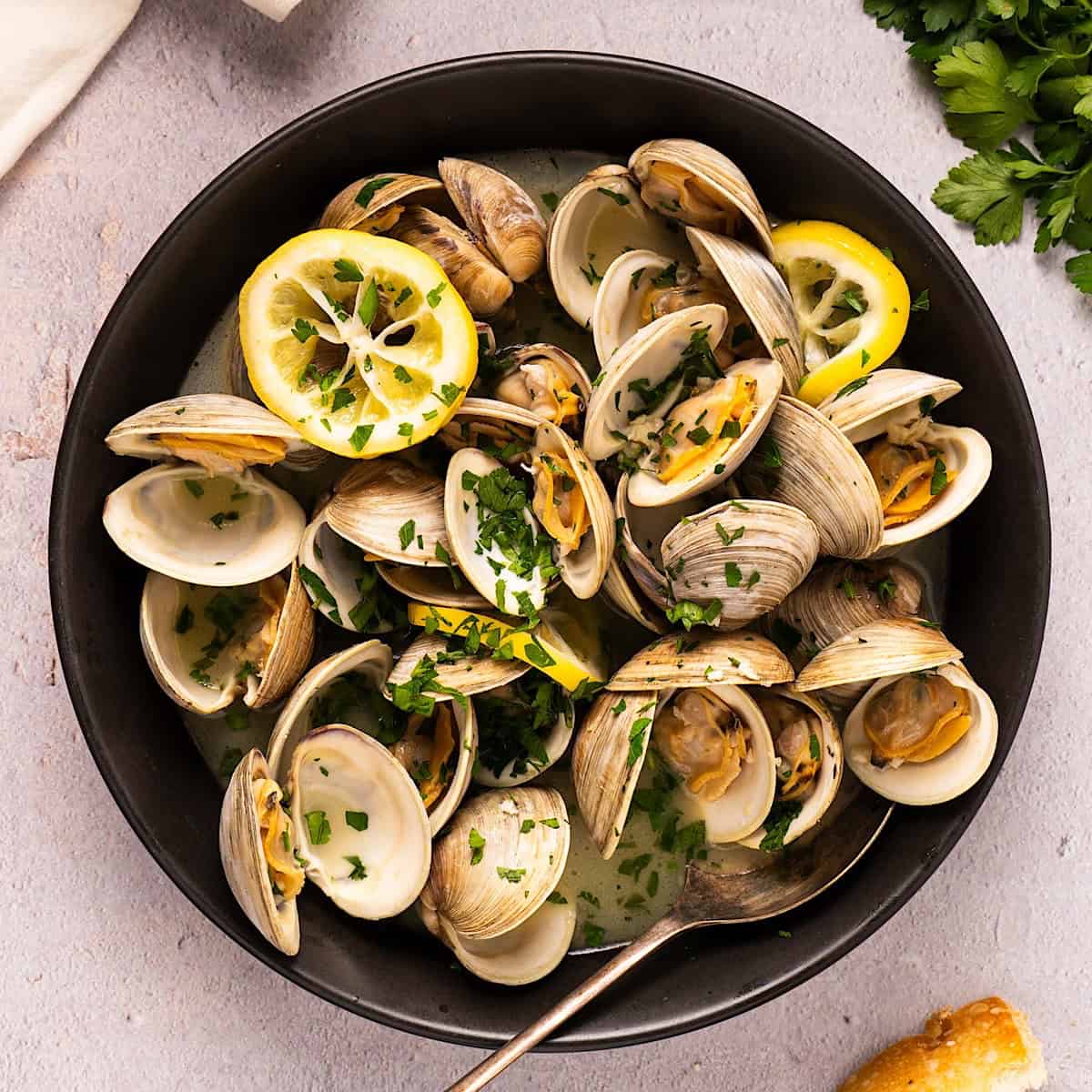 Easy steamed littleneck clams in a black bowl plated next to linen, parsley, and bread.