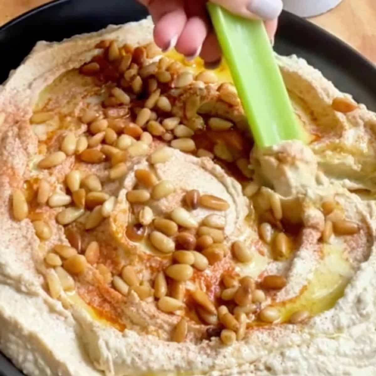 Dipping celery in Greek hummus that has been transferred to a serving bowl with pine nuts.
