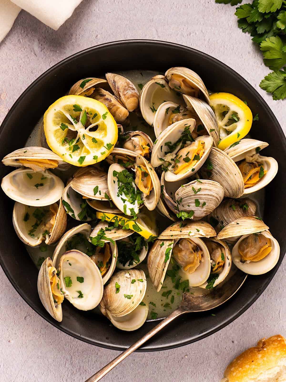 Easy steamed littleneck clams in a black bowl plated next to linen, parsley, and bread.