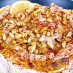 Authentic greek hummus recipe with lemon garlic and pine nuts feature image close up in large serving bowl with topping and lemon wedge.