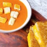 Roasted tomato basil soup with croutons and sourdough grilled cheese on a wooden platter.
