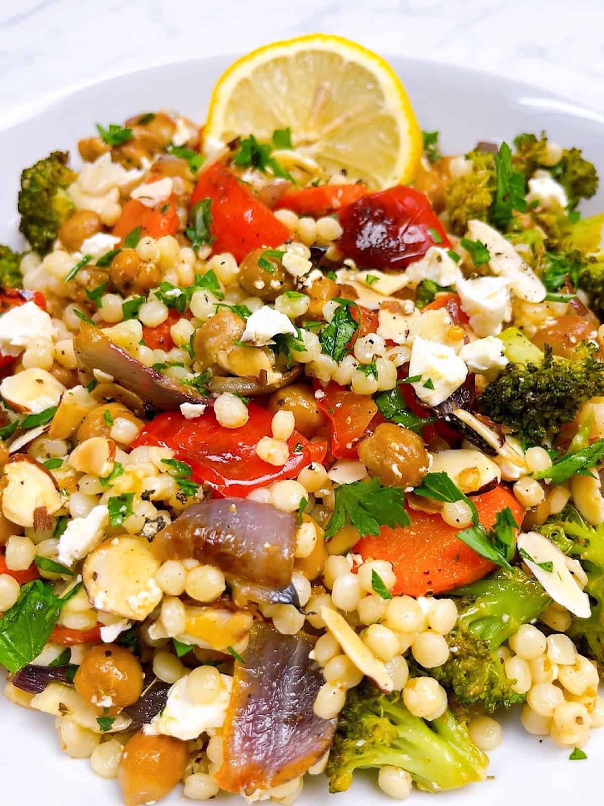 Couscous with roasted veggies, chickpeas, and feta hero image close up in bowl with lemon garnish.