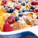 Brioche french toast bake with eggnog feature image close up in casserole dish baked with drizzle topping, berries, and powdered sugar.