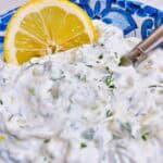 Tzatziki sauce in a blue bowl with decorative utensil and lemon wedge.
