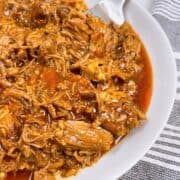 Pulled pork bbq with juices in white bowl with a large silver fork and cloth towel.