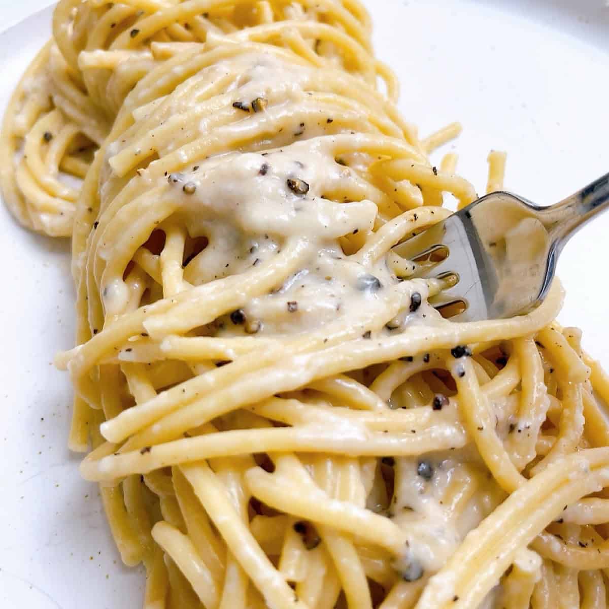 Bucatini cacio e pepe twirled on a plate with pepper sauce on top and a fork inserted.