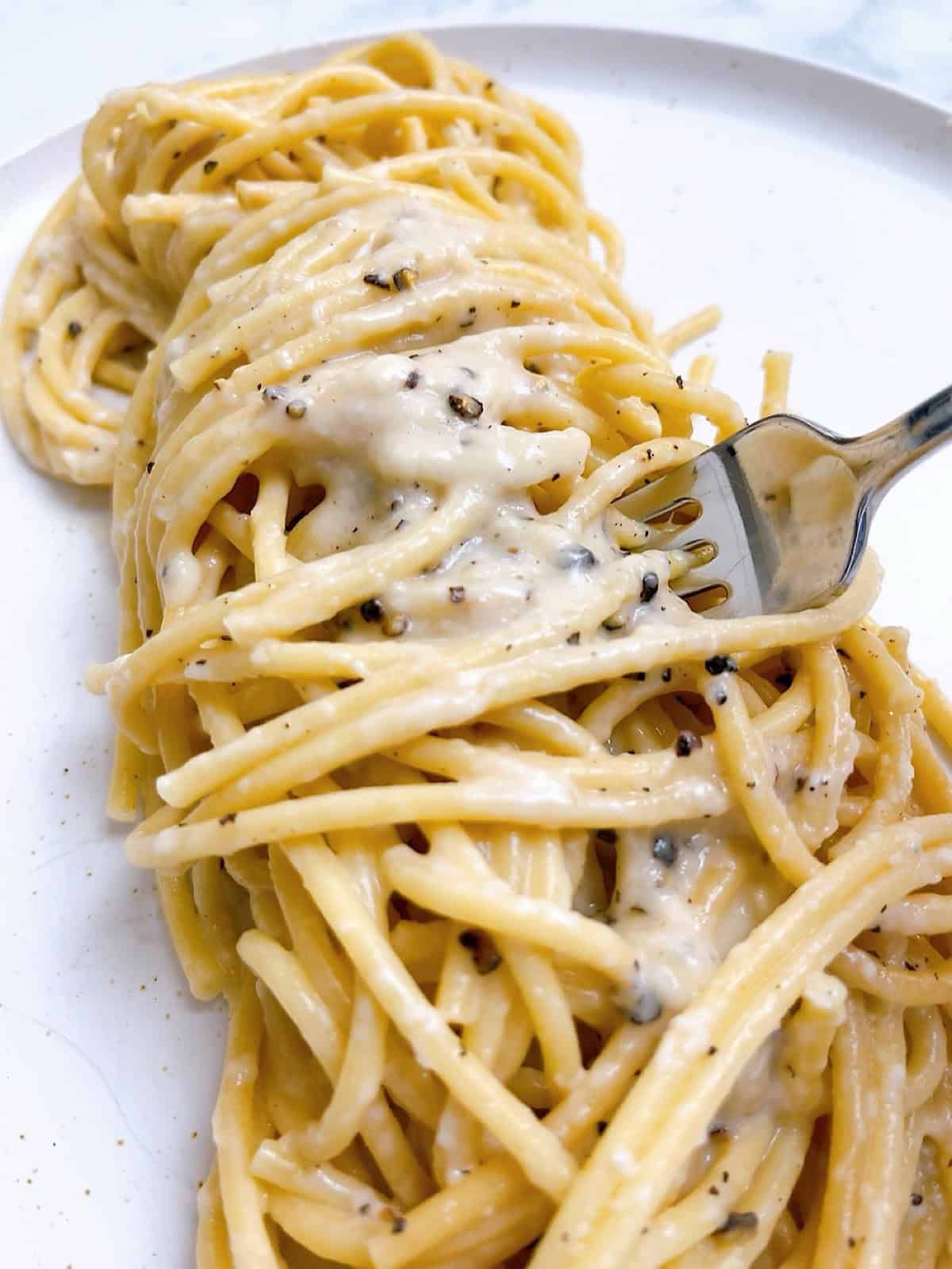 Bucatini cacio e pepe twirled on a plate with pepper sauce on top and a fork inserted.