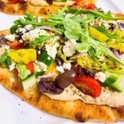Two Mediterranean flatbread pizzas on parchment with all the toppings.