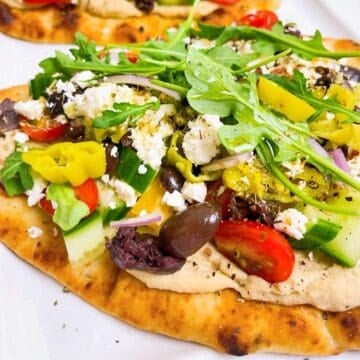 Two Mediterranean flatbread pizzas on parchment with all the toppings.