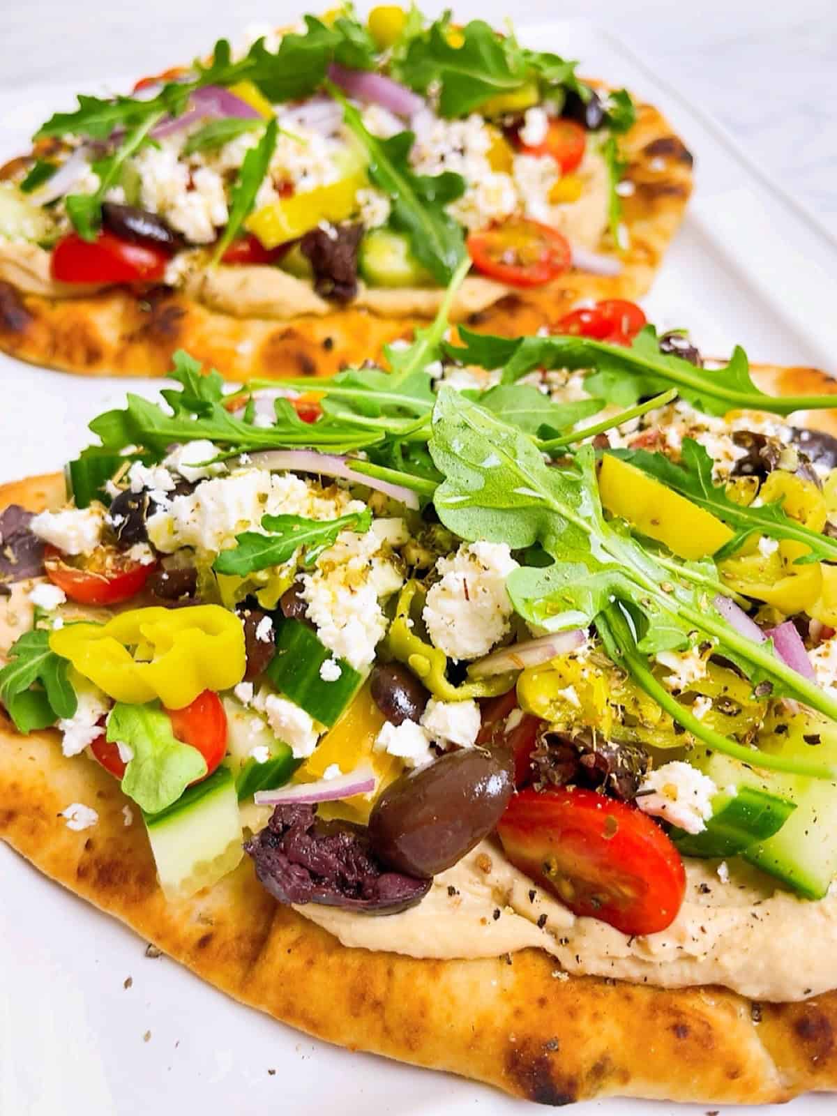 Two Mediterranean flatbread pizzas on parchment with all the toppings.