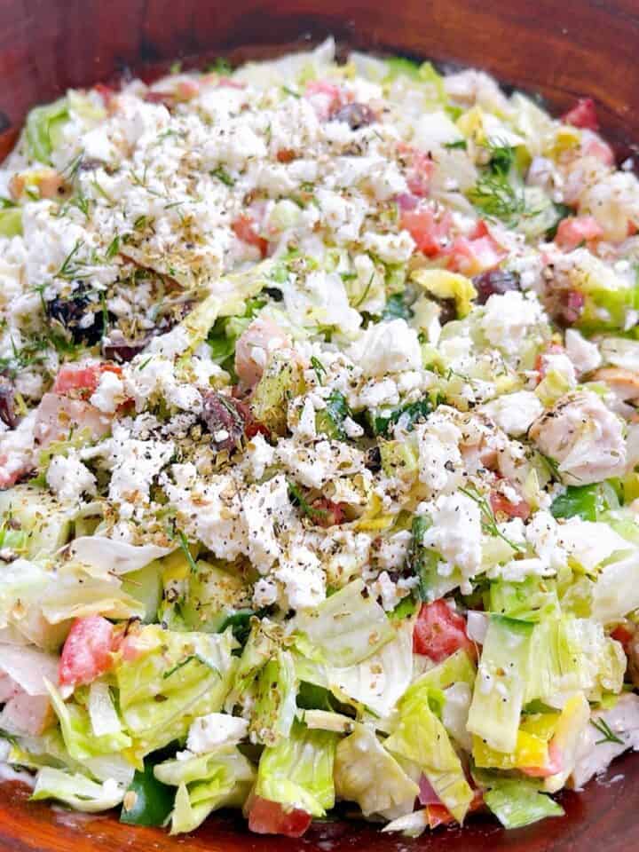 A tossed chopped Greek salad in wooden salad bowl topped with feta, dill, and oregano.