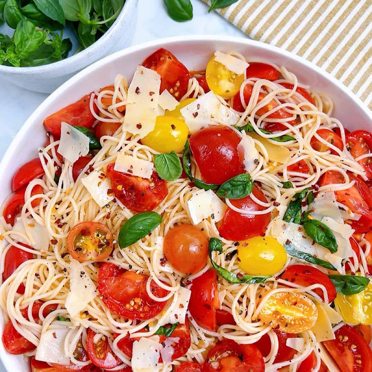 Spaghetti salad in large white bowl with garnish and a bowl of basil off to the side.