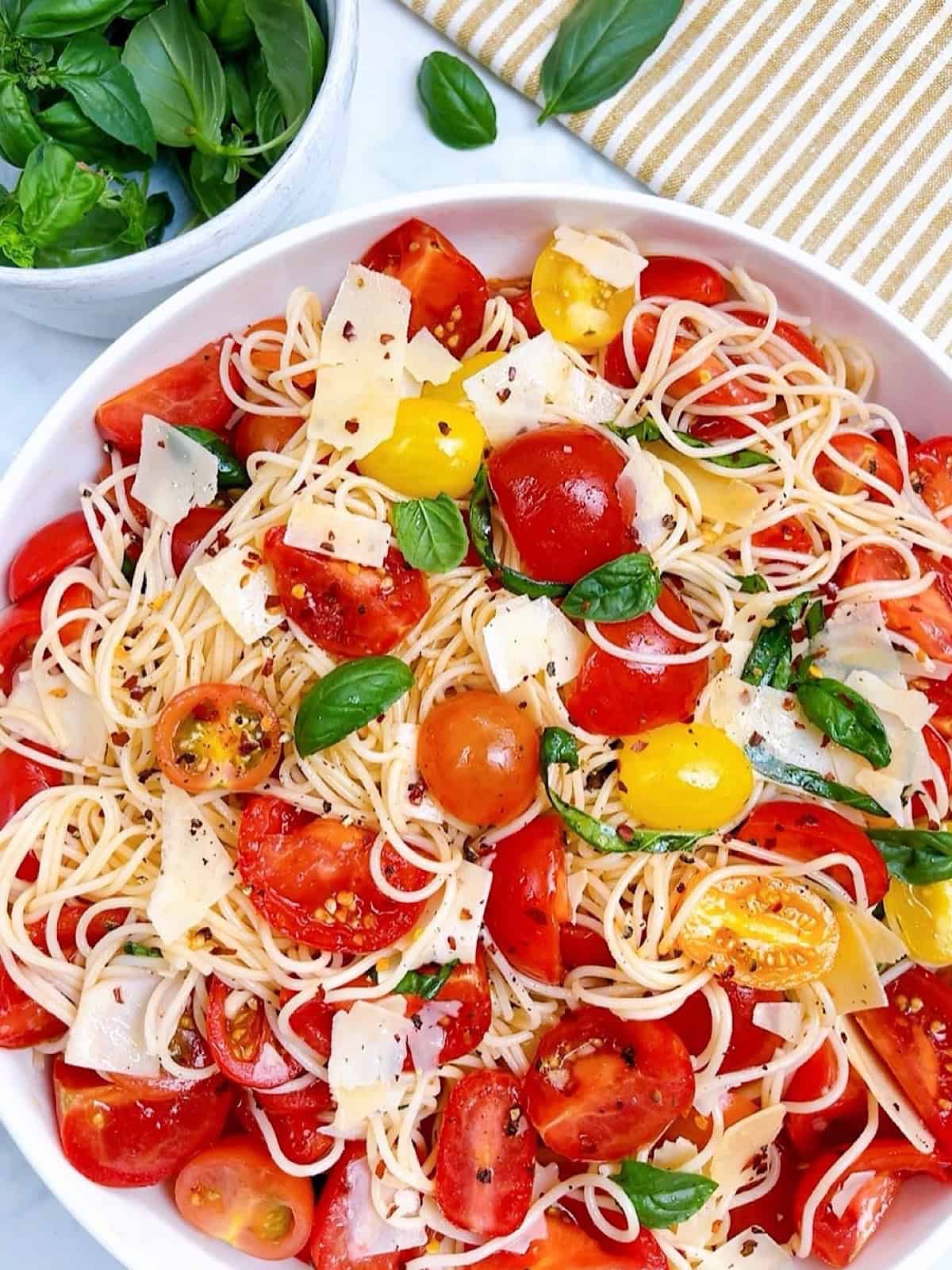 Spaghetti salad in large white bowl with garnish and a bowl of basil off to the side.