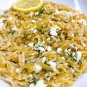 Creamy lemon orzo pasta in a white bowl with dill, feta, and lemon as garnish.