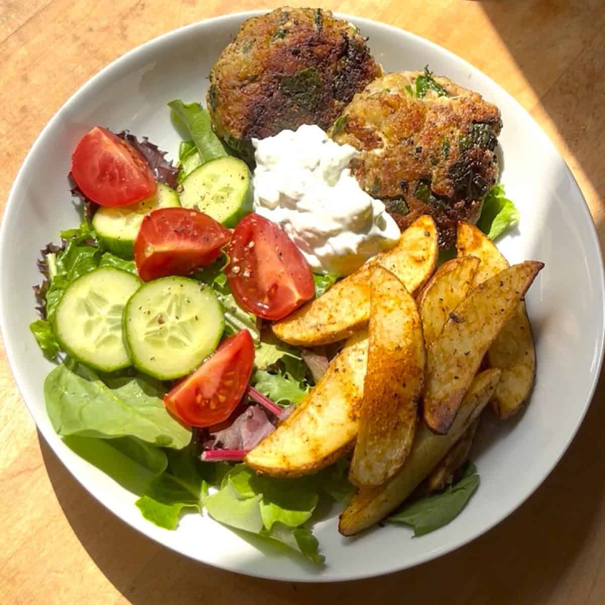 Cooked turkey burgers nestled in a bowl on a bed of greens with tomato, cucumber, potato wedges, and tzatziki sauce.