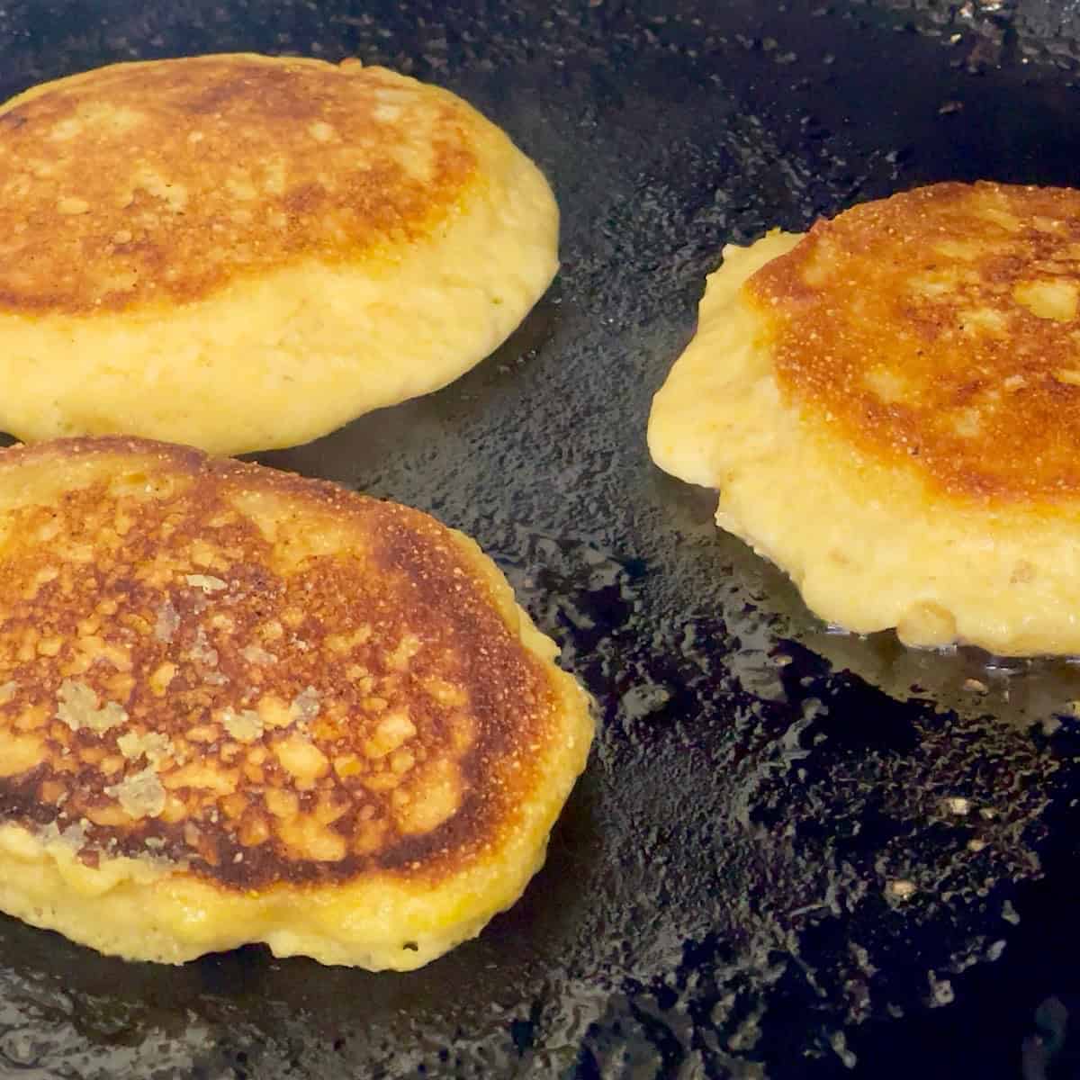 Three hoecakes cooking in a cast iron skillet.
