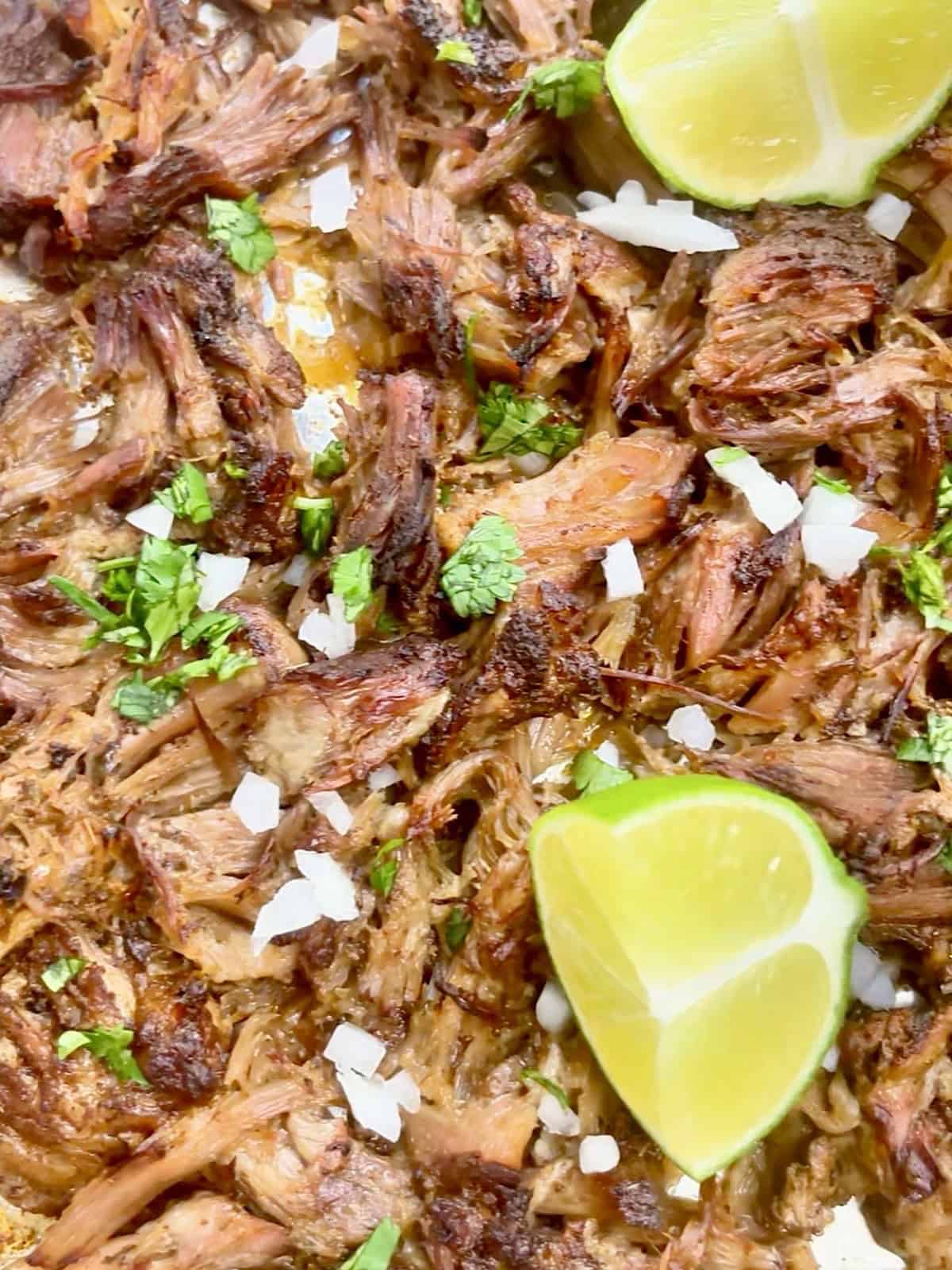 Mexican pulled pork with crispy edges on a sheet pan topped with onion, cilantro, and lime wedges.