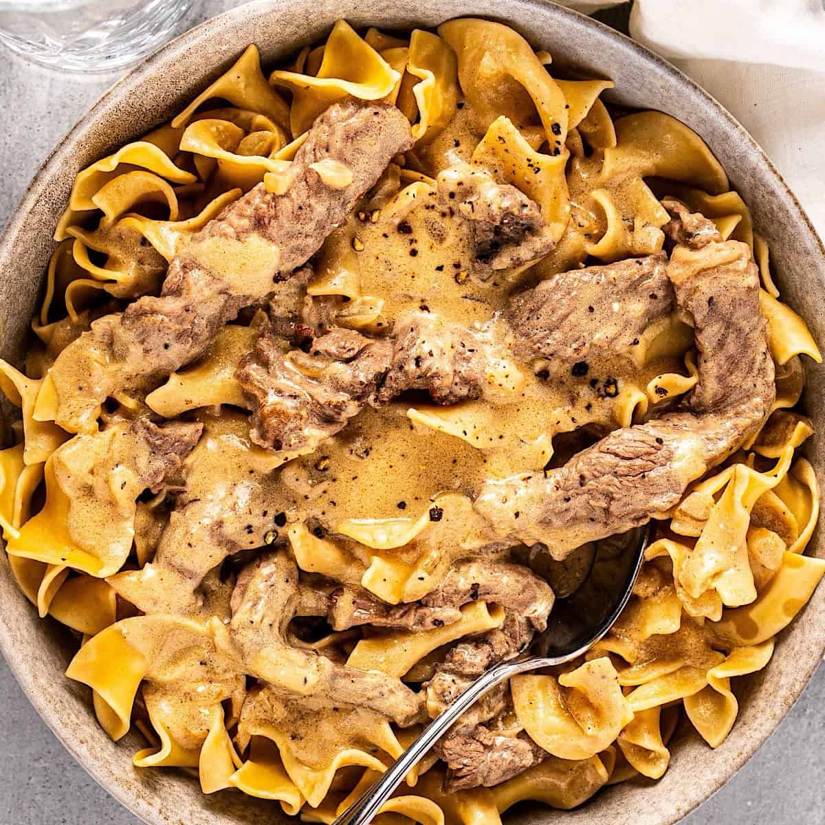 A bowl of beef stroganoff with a spoon inside and a linen napkin nearby.